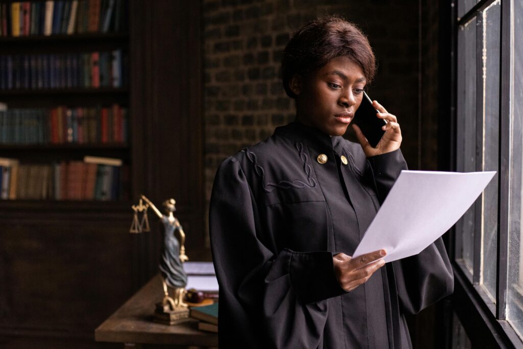African American judge in robe, reading document and talking on phone by the office window.
